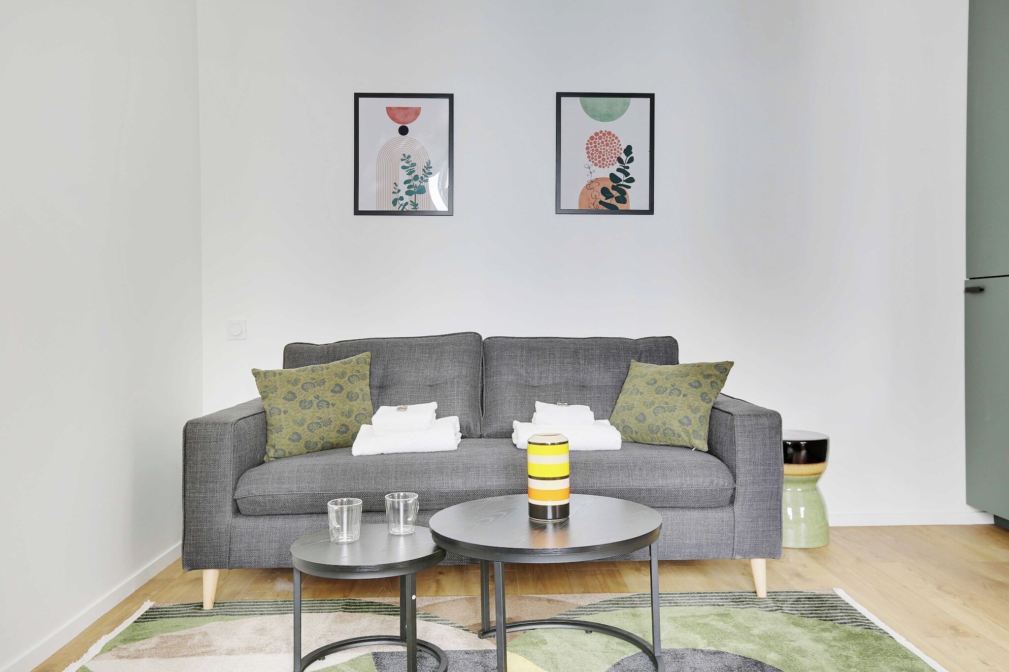 A living room with a grey sofa, two side tables, a yellow and black thermos, and two framed artworks on the wall.