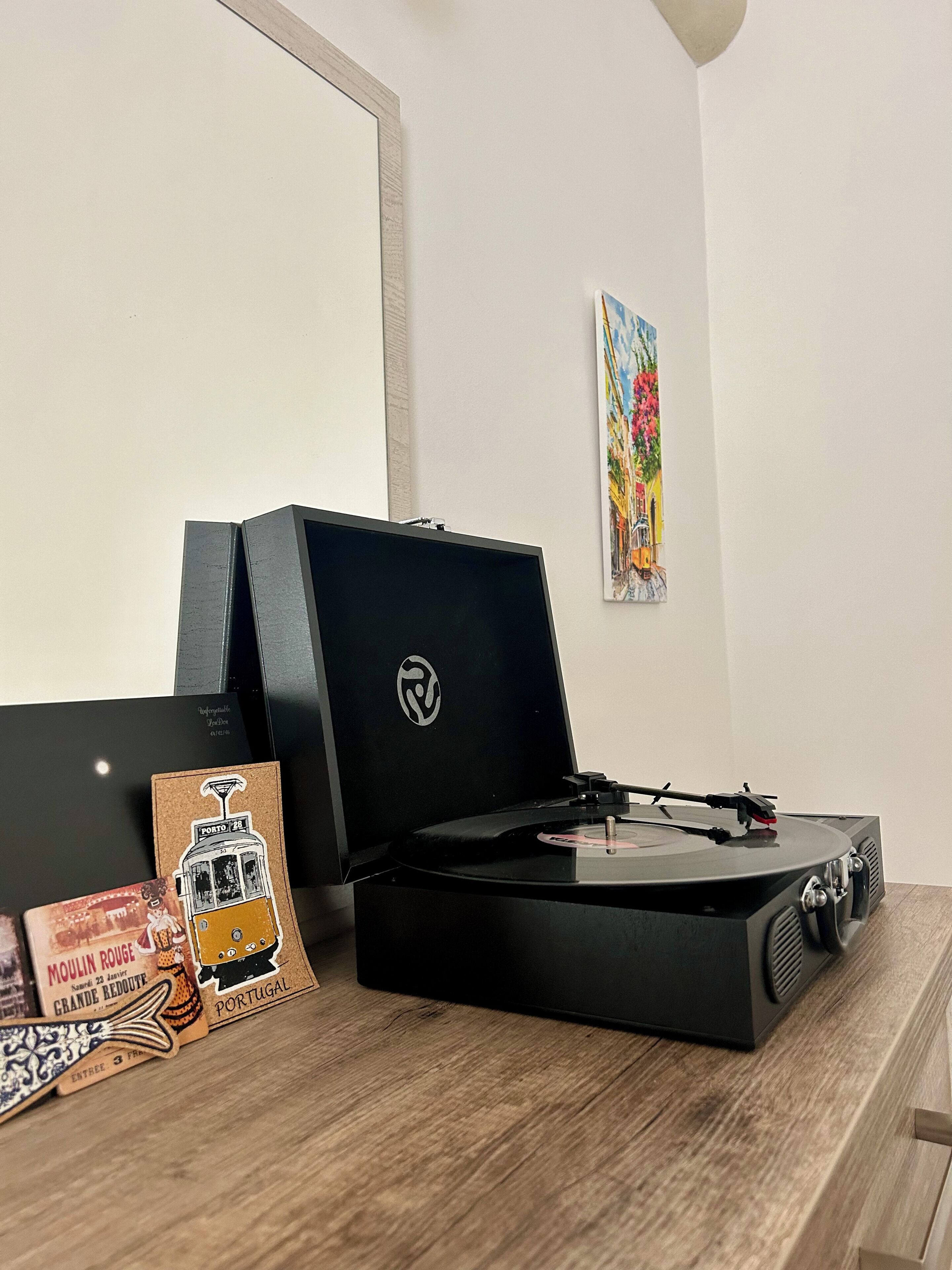 A turntable on a wooden surface with a vintage postcard and a colorful painting in the background.