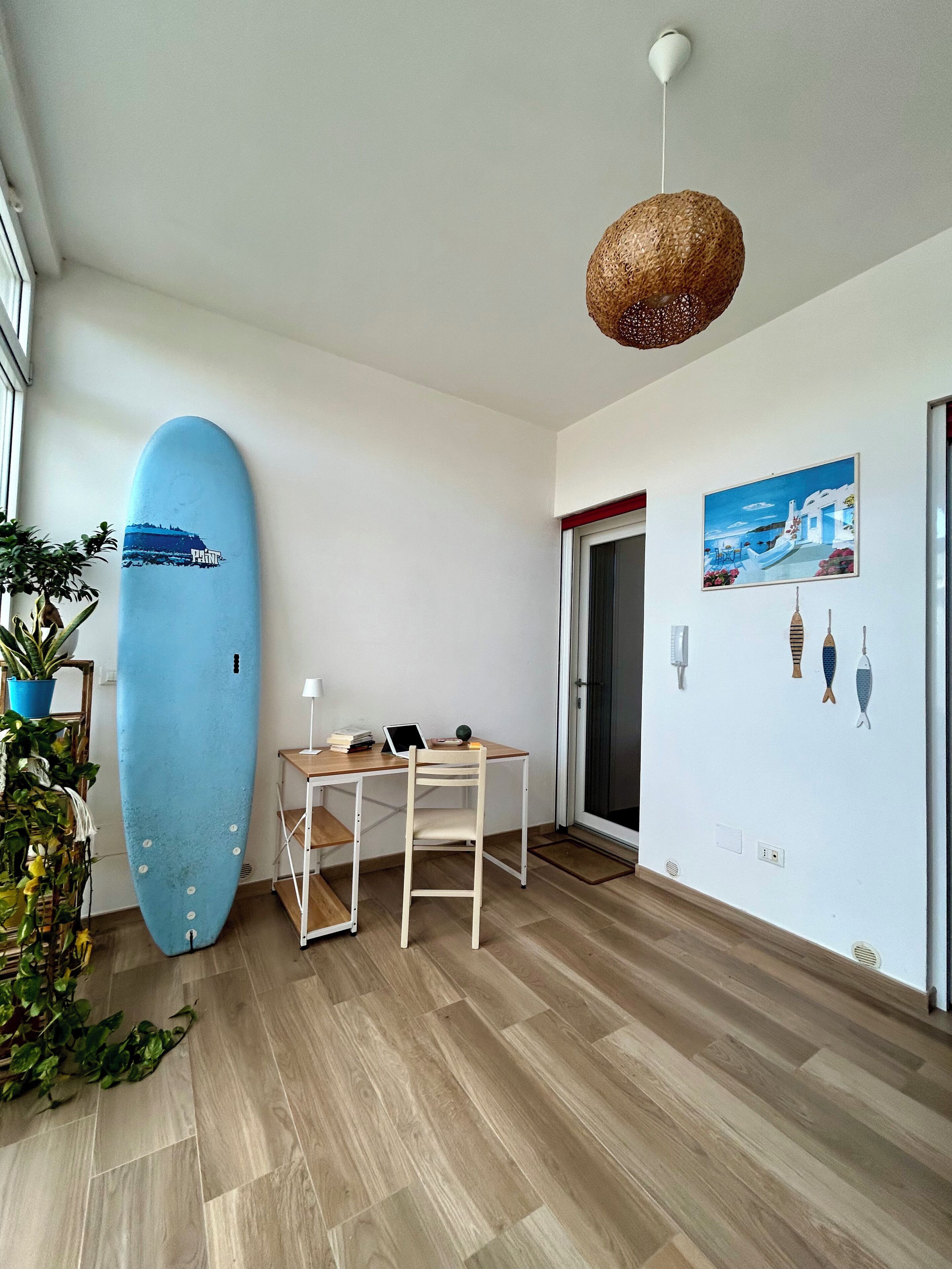 A room with a wooden floor, a desk with a chair, a surfboard, a hanging light fixture, and a wall with fish illustrations.