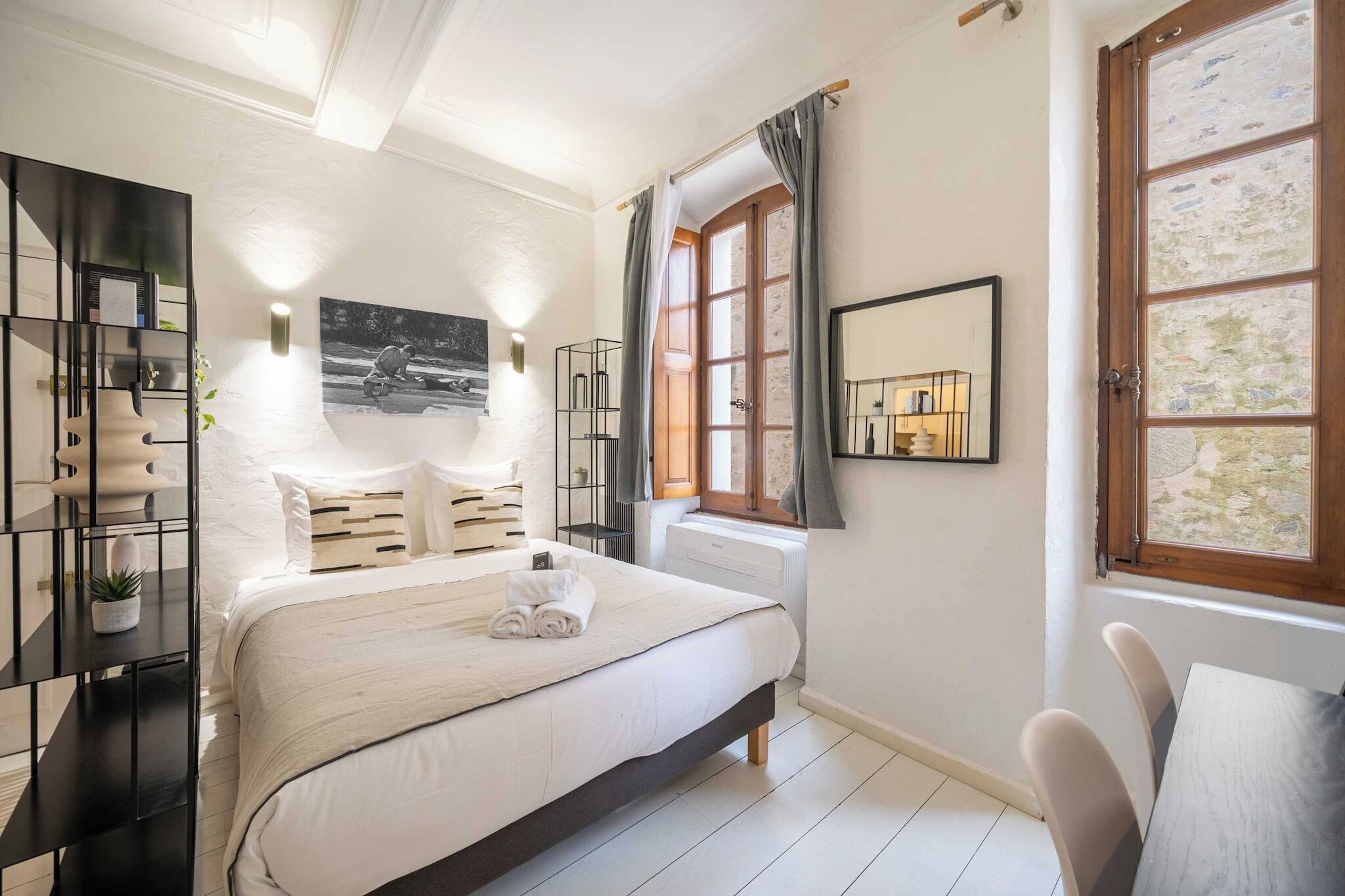 Ein Schlafzimmer mit einem Bett, einem Nachttisch, einem Heizkörper, einem Fenster mit Vorhängen und einem an der Wand befestigten Spiegel.