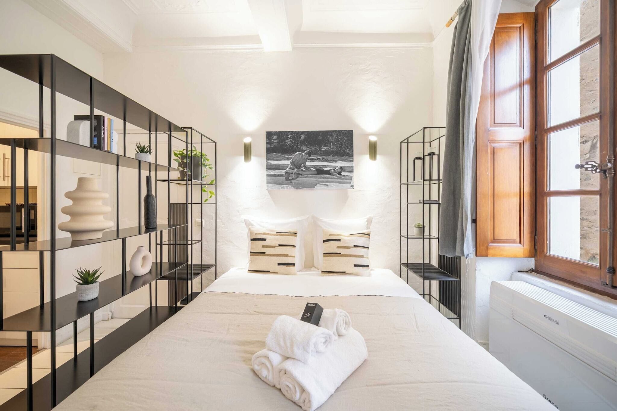 A modern hotel room with a large bed, two bedside tables, a black metal shelving unit, a window with curtains, and a wall-mounted picture.