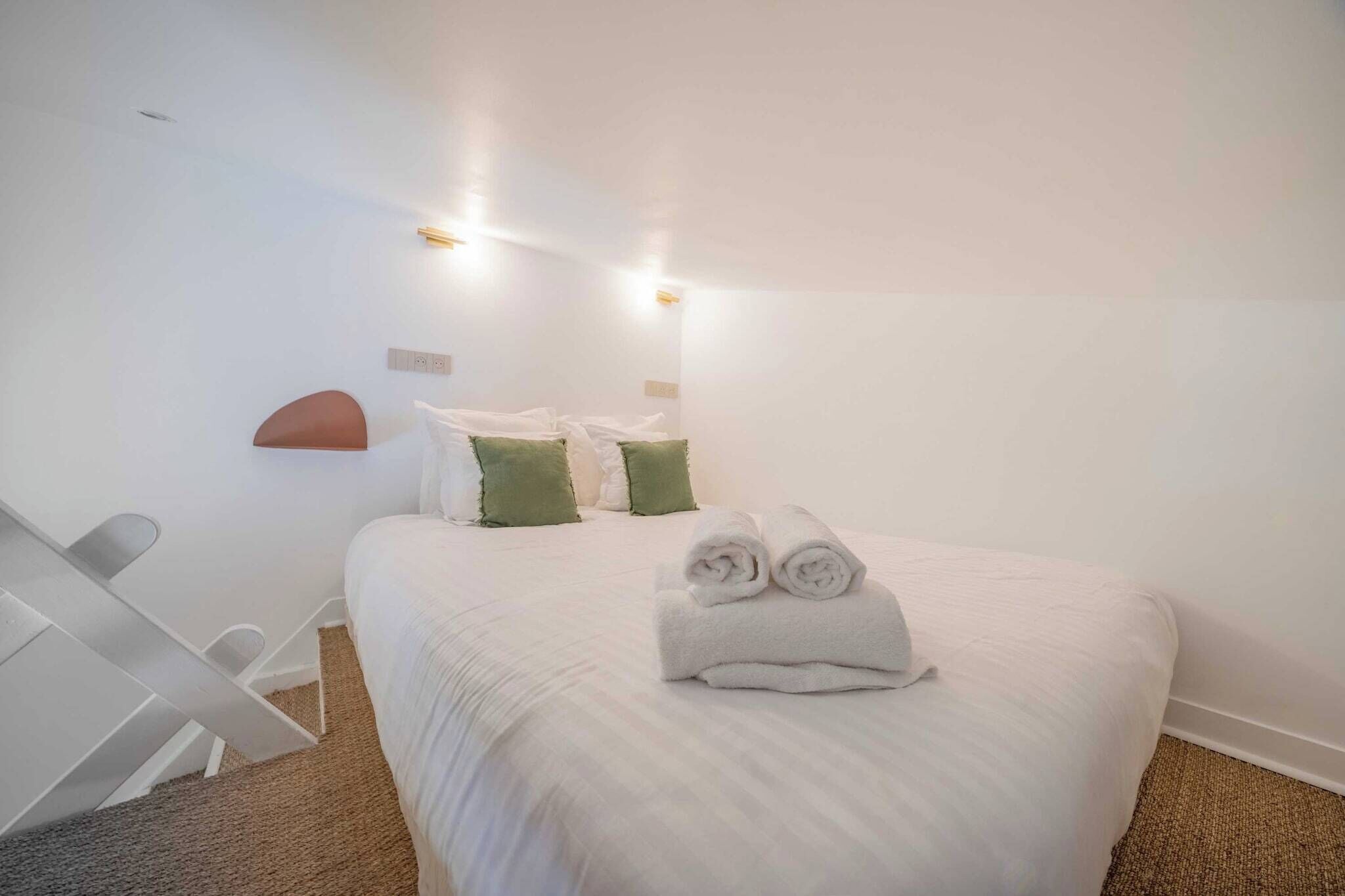 A hotel room with a bed, two pillows, a rolled towel, a white chair, and a wall-mounted light fixture.