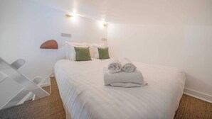 Habitación de hotel con cama, dos almohadas, toalla enrollada, una silla blanca y una lámpara fijada a la pared.