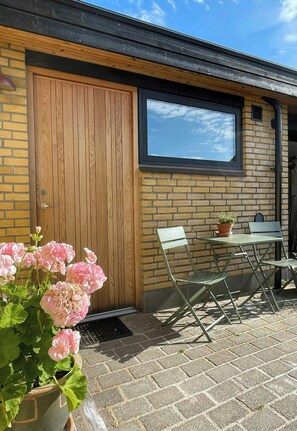 A wooden door with a window above it, a brick wall, a small table with a chair, and a potted plant with pink flowers. - Retro 70s house with lake views in Jelling (Jelling)
