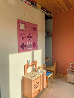 Ein Kinderzimmer mit einer rosa Decke, Holzsesseln, einem Spielherd und einem Bücherregal voller Bücher.