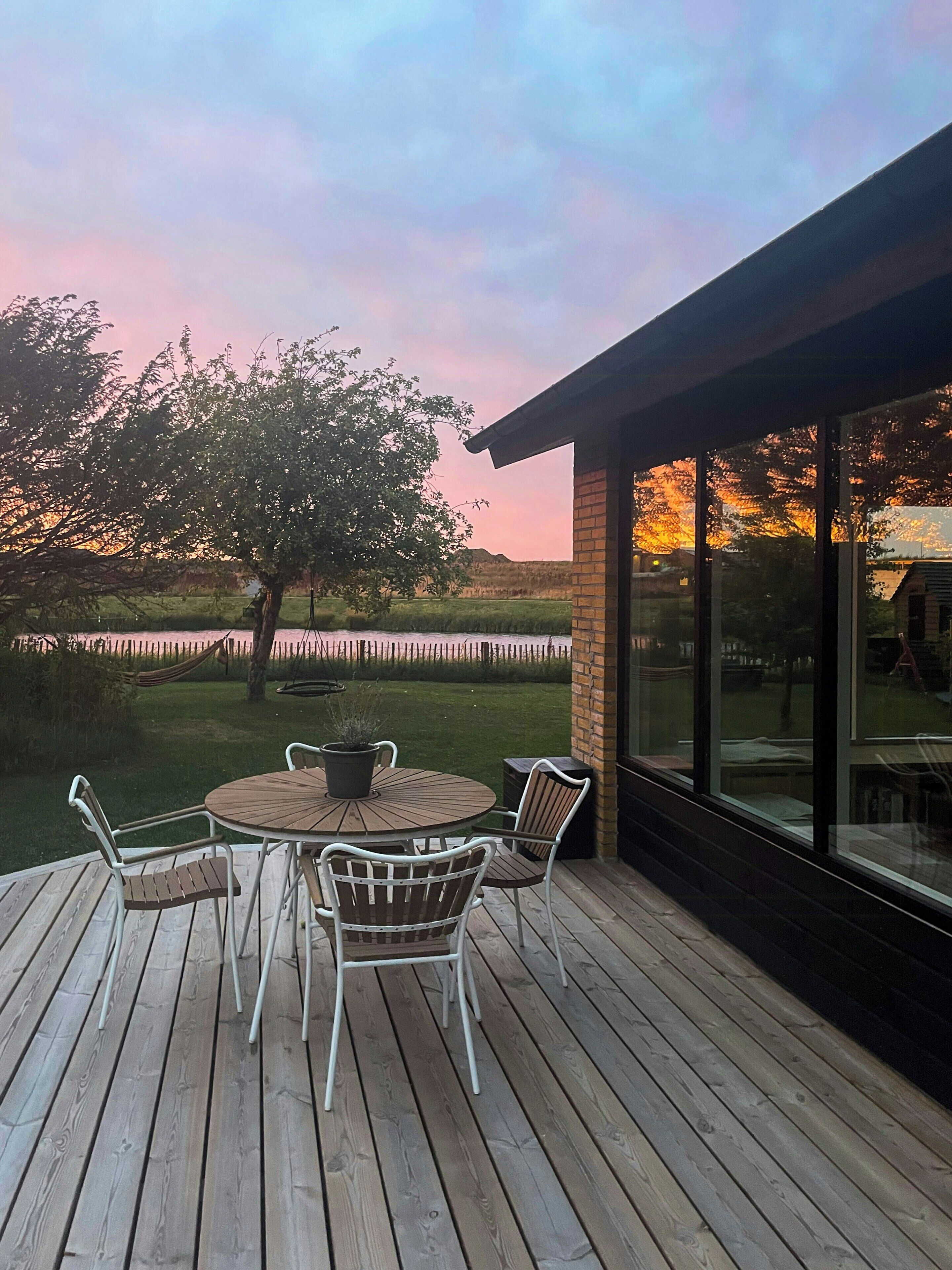 Una terraza de madera con una mesa redonda y sillas, con vista a un cuerpo de agua y un área verde con un árbol.
