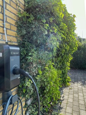 A black electric vehicle charging station labeled 'Clever' with a cable plugged into it, positioned against a brick wall with ivy growing over it. - Retro 70s house with lake views in Jelling (Jelling)