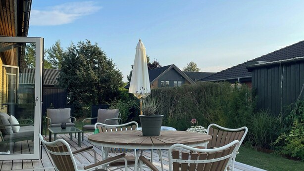 En udendørs terrasse med et rundt bord og stole, en potteplante og en hvid parasol.