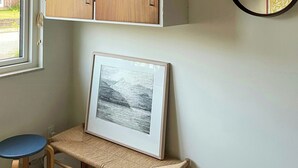 Et værelse med træloft, et indrammet billede på en hylde, et rundt spejl på væggen og et vindue med persienner.