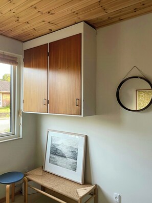 A room with a wooden ceiling, a framed picture on a shelf, a round mirror on the wall, and a window with blinds.