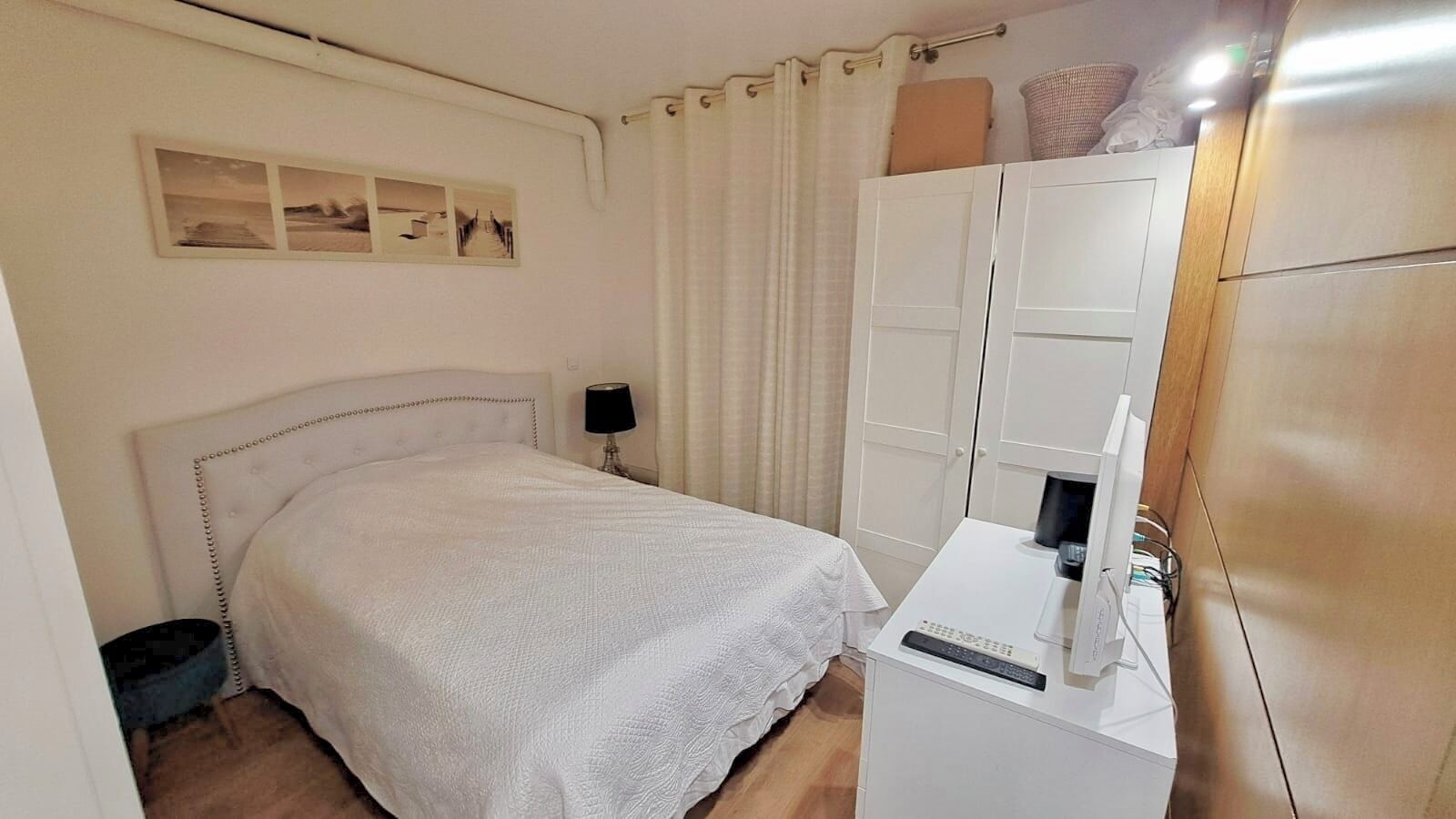 A bedroom with a bed, a nightstand, a wardrobe, and a desk with a computer and a lamp.