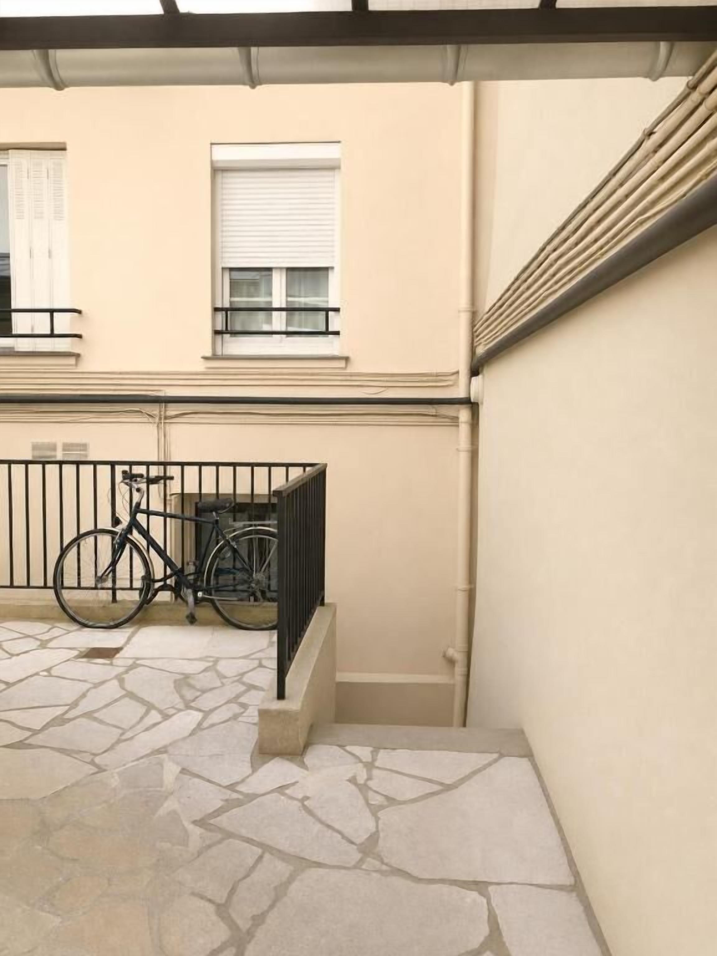 A stone-paved courtyard with a bicycle, a metal railing, and a building with windows and a door.
