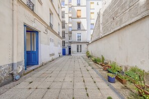 A narrow alleyway with a blue door on the left, potted plants along the right, and a building with multiple windows in the background. - Design Studio for 2P – Batignolles (Paris)