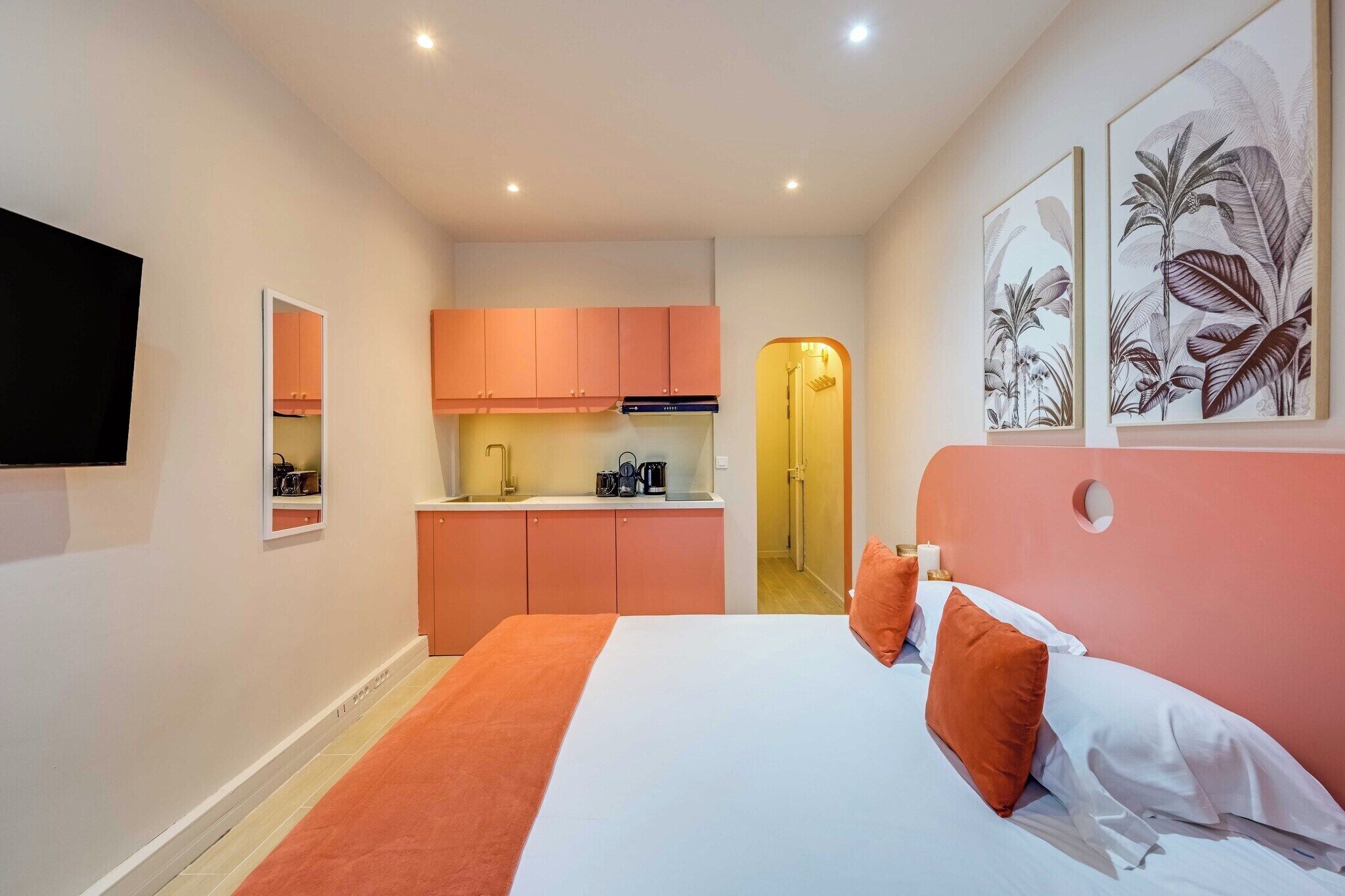 A compact hotel room with a bed, a kitchenette, and a TV mounted on the wall.