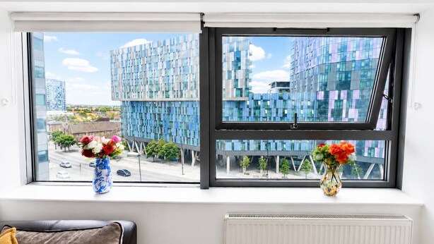 A room with a large window overlooking modern buildings, a radiator, and two vases with flowers. - Spacious 2BR Sleeps 7,  Long Stays (Greater London)