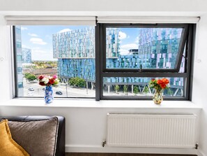 A room with a large window overlooking modern buildings, a radiator below, and two vases with flowers on the windowsill. - Spacious 2BR Sleeps 7,  Long Stays (Greater London)