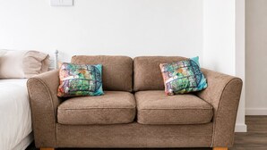 A brown sofa with two colorful pillows, a white wall, and a bed with a white pillow. - Spacious 2BR Sleeps 7,  Long Stays (Greater London)