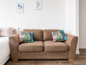 A brown sofa with two colorful pillows, a white wall, and a bed with a white pillow. - Spacious 2BR Sleeps 7,  Long Stays (Greater London)