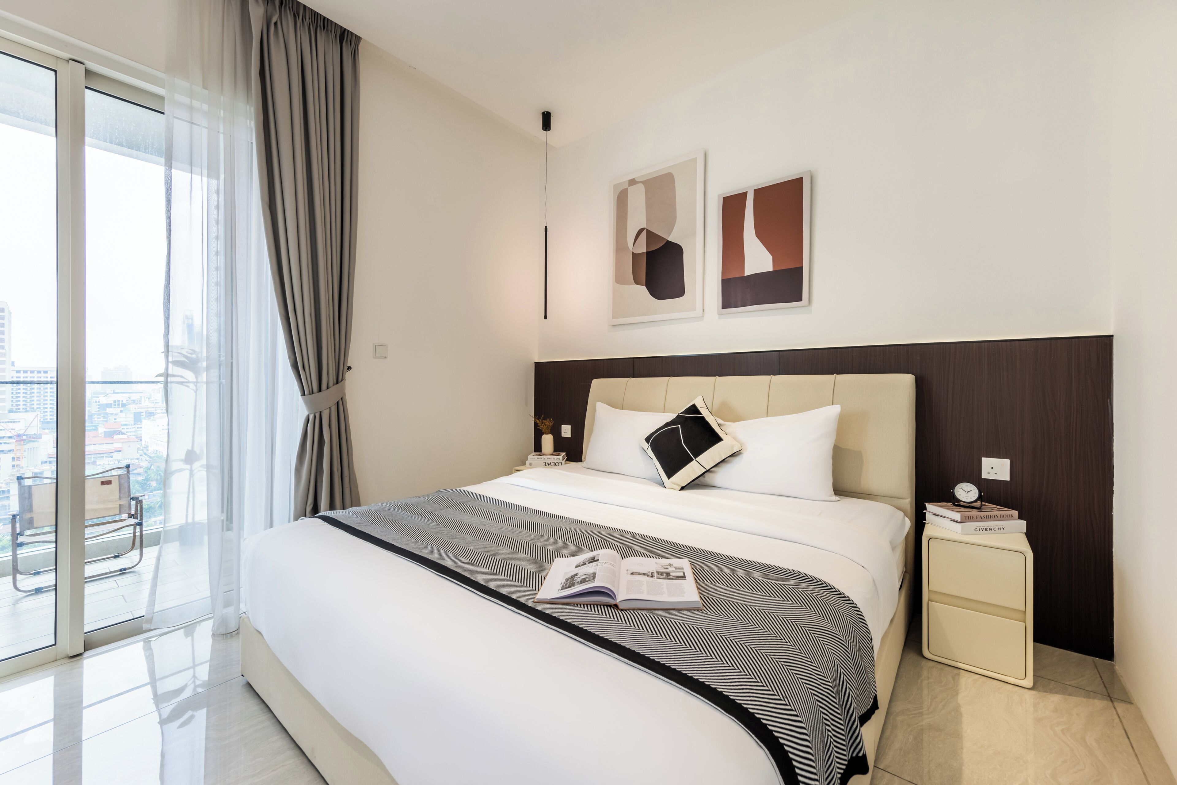 A hotel room with a large bed, a bedside table, a chair, and a view of the city through the window.