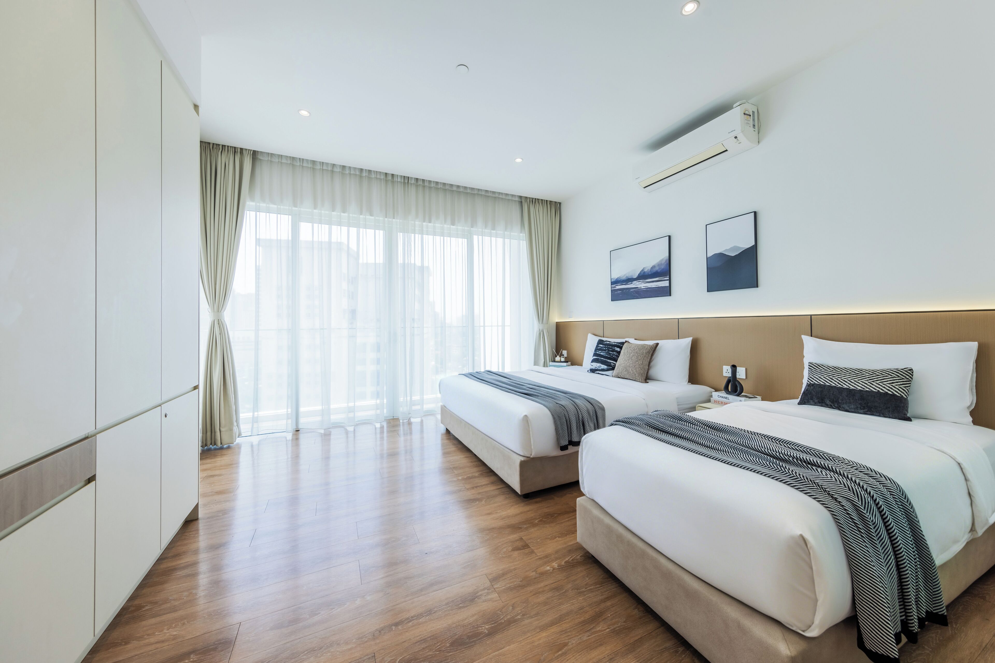 A hotel room with two beds, a large window with sheer curtains, a wall-mounted air conditioner, and a framed picture.