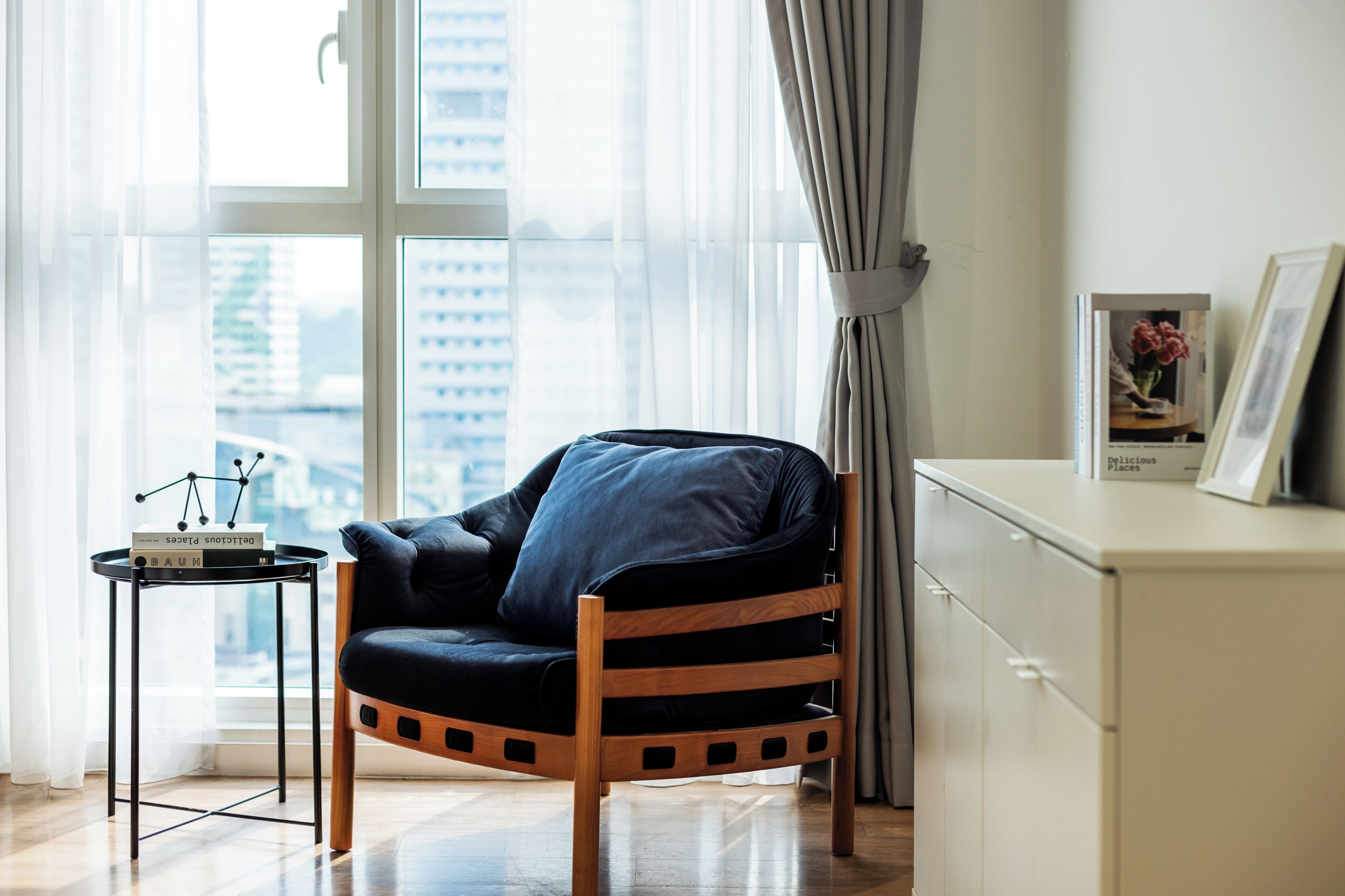 Una sala moderna con un sillón de cuero negro, una mesita de noche con libros, un armario blanco y vistas a la ciudad a través de amplias ventanas.