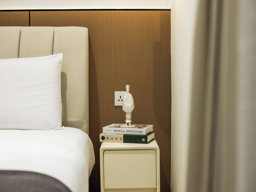 A neatly made bed with white bedding, a bedside table with a lamp and books, and a wooden headboard.