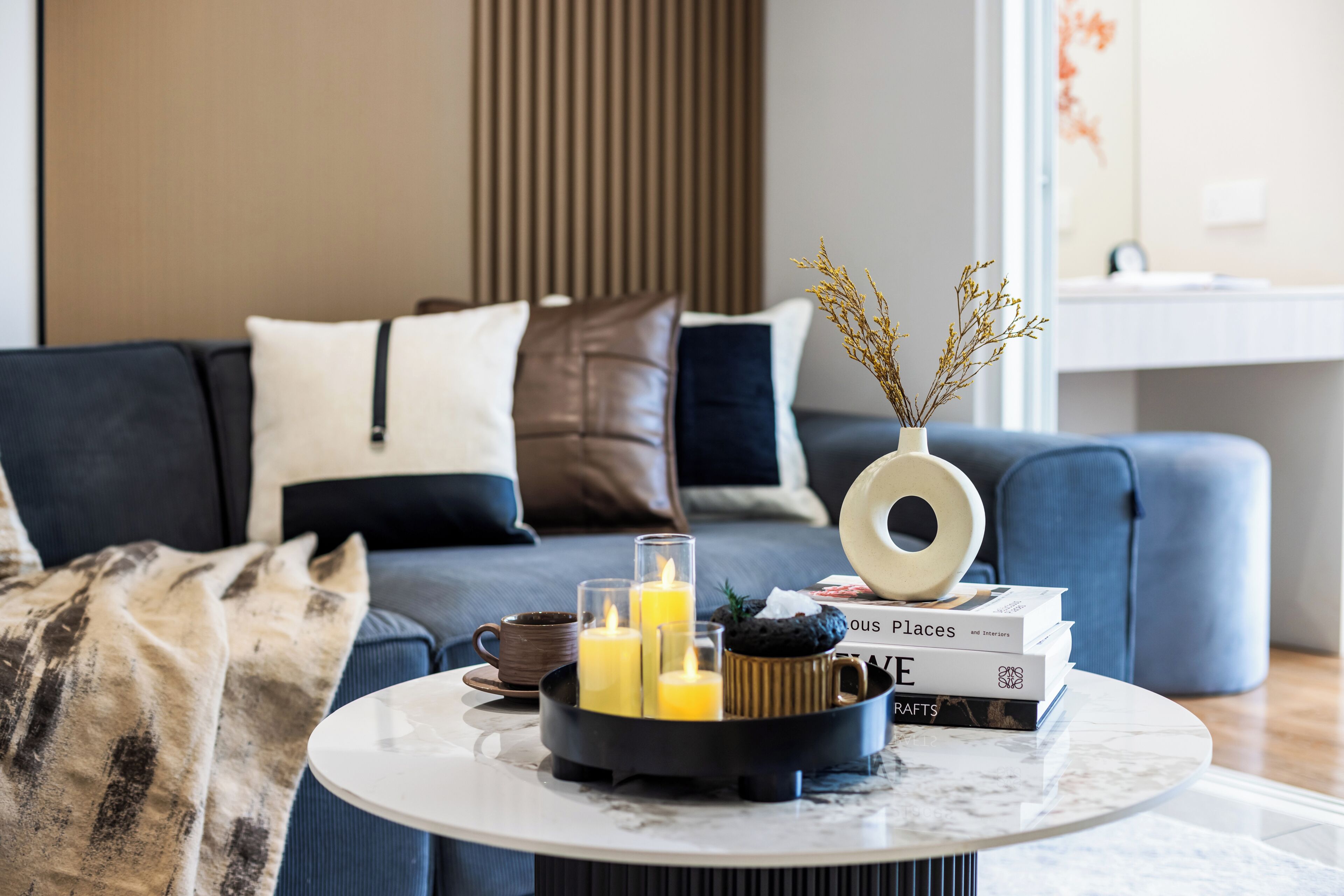 A modern living room with a sofa, a coffee table with books and candles, and a vase with dried plants.
