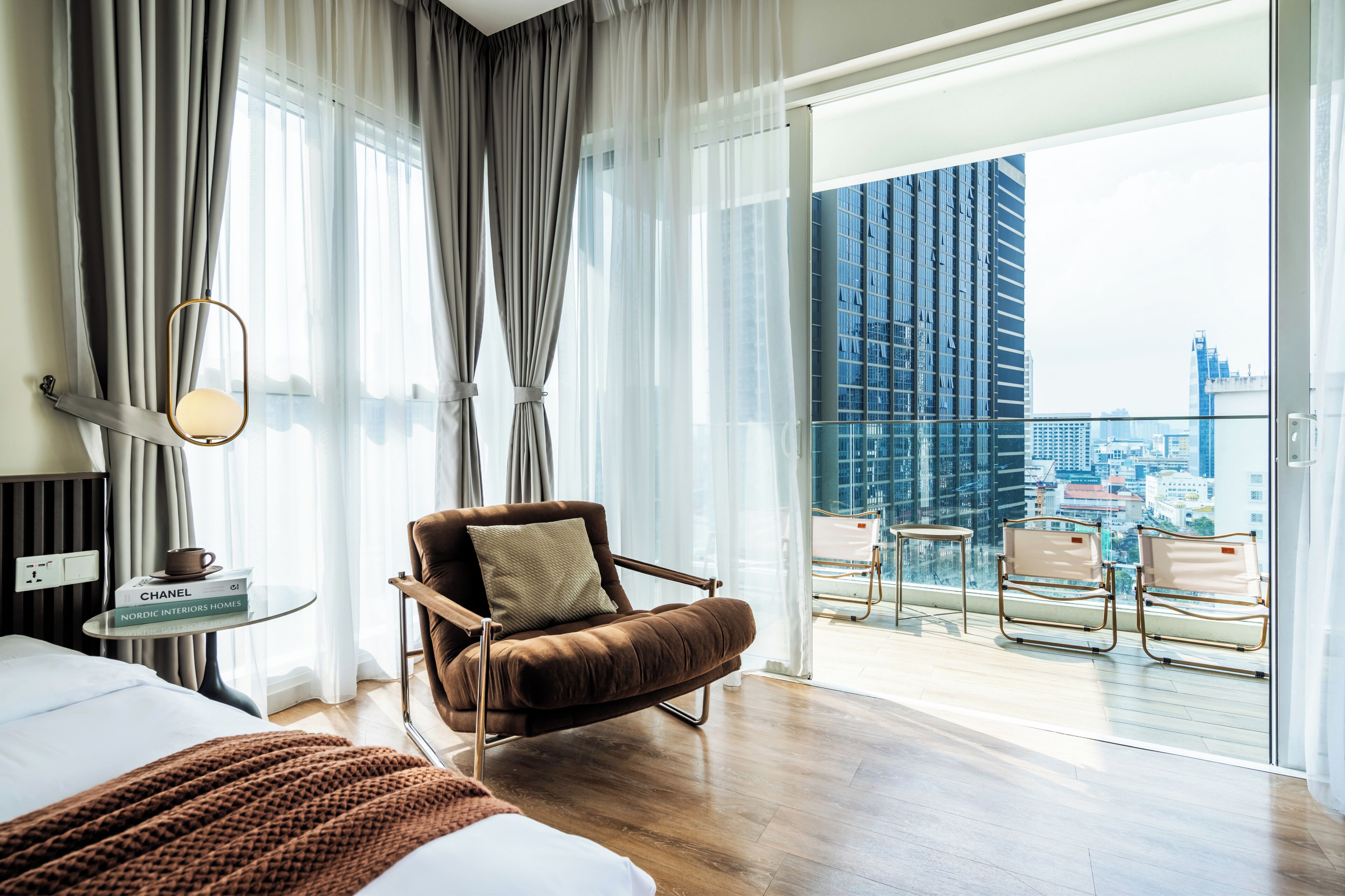 A hotel room with a bed, a brown armchair, a small table with books, and a view of a cityscape through large windows.