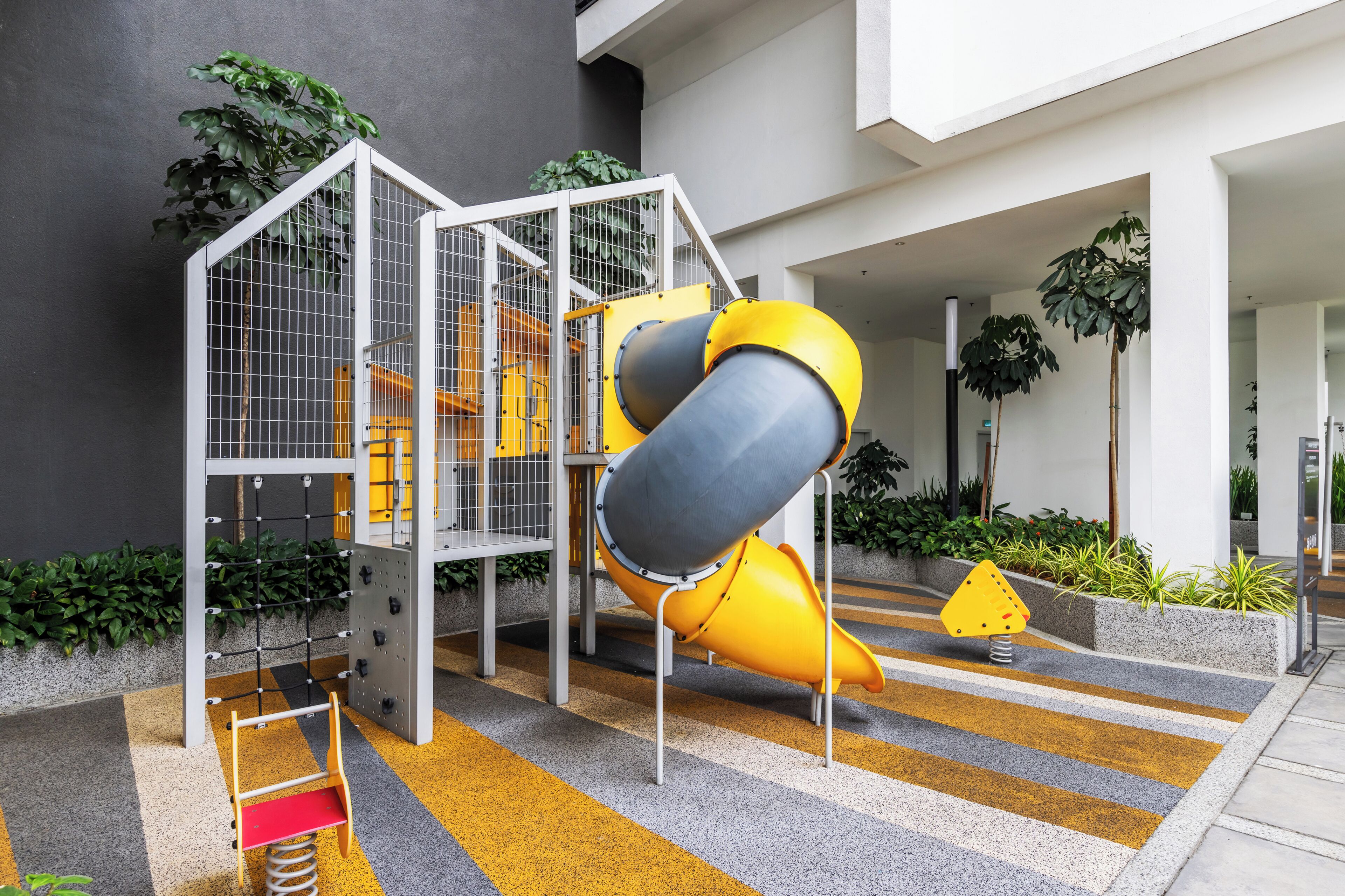 A playground with a slide and climbing structures, surrounded by plants and a modern building facade.