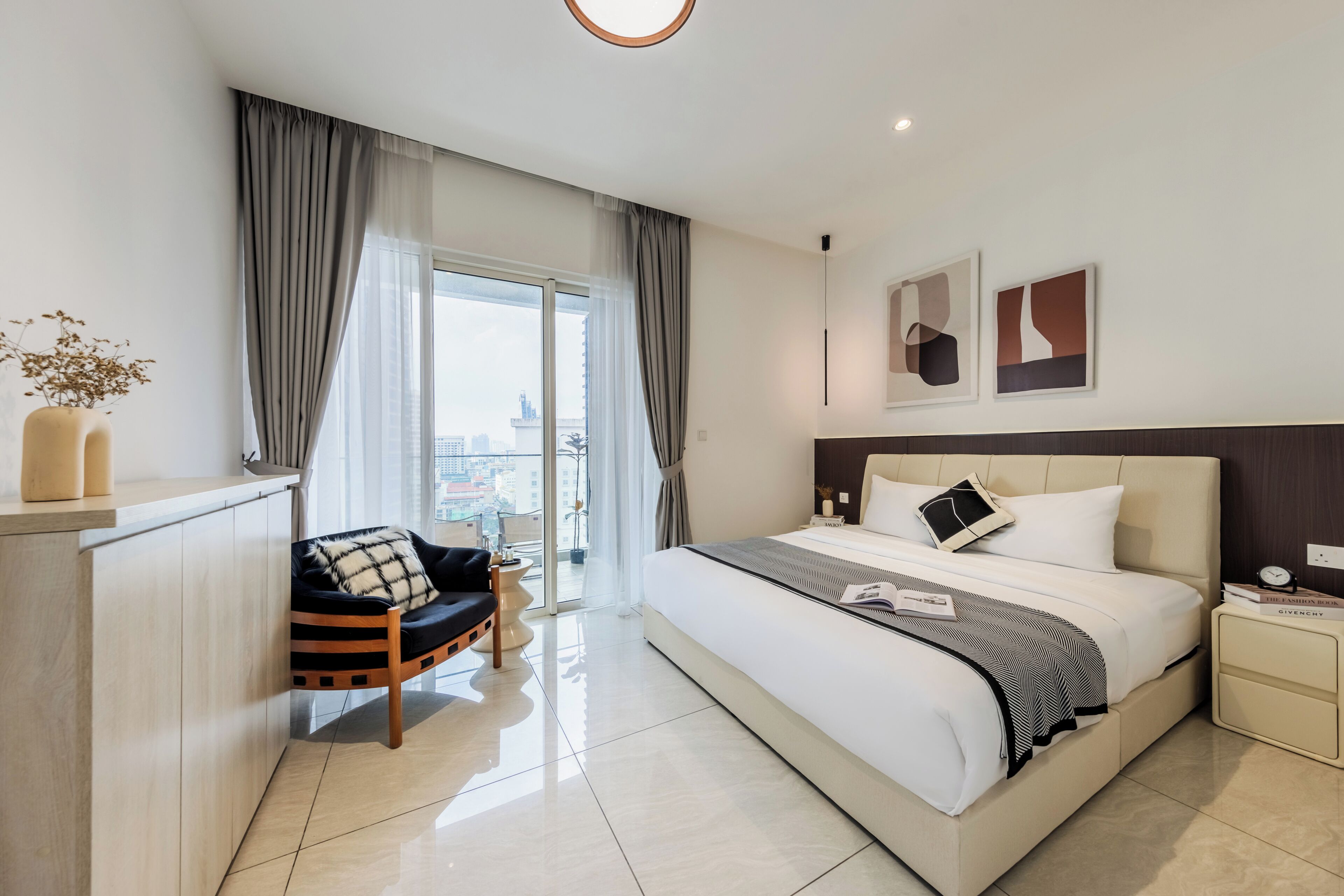 A modern hotel room with a large bed, a seating area with a chair, a bedside table with books, and a view of the city through the window.