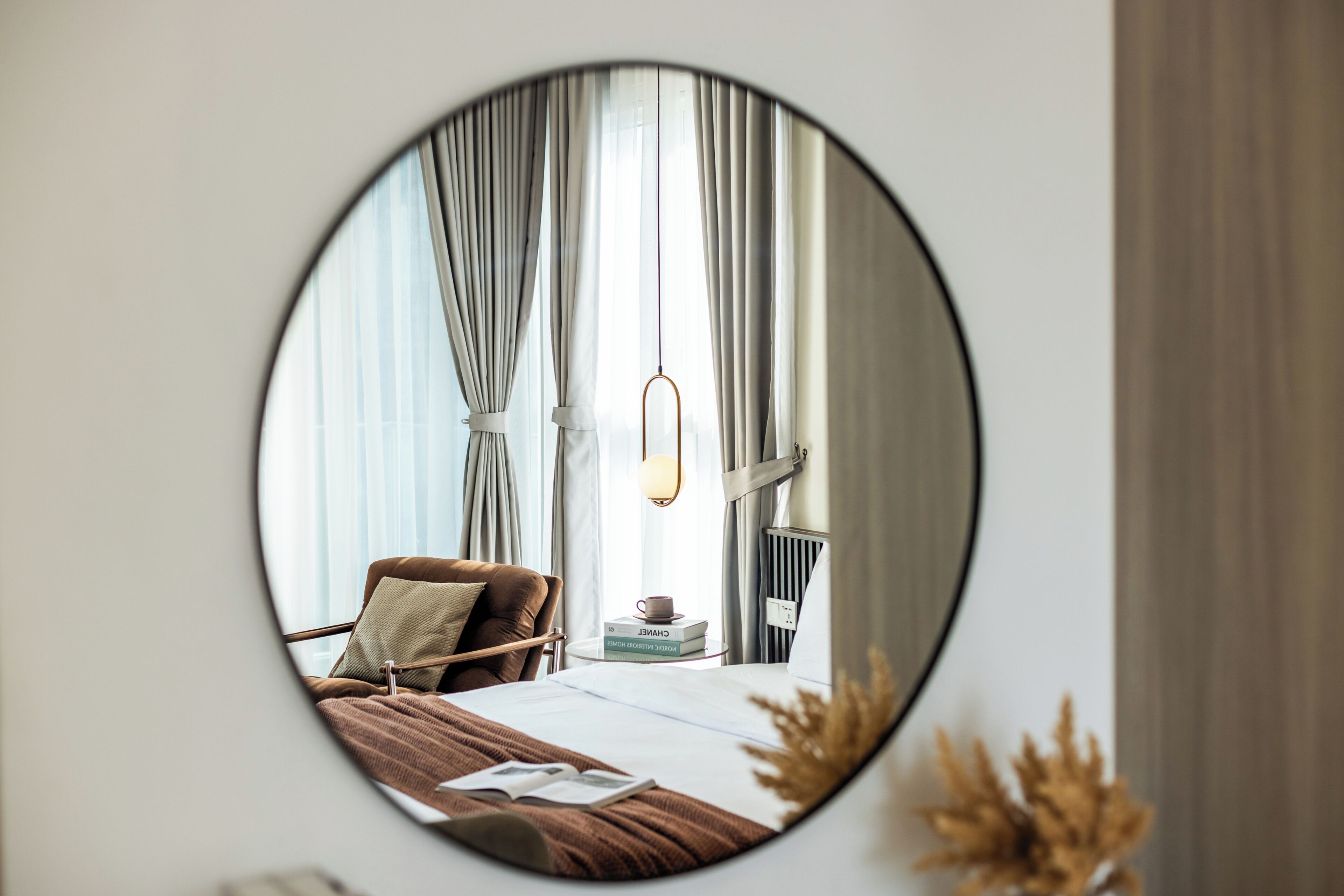 A round mirror reflecting a bedroom with a bed, pillows, a bedside table with books, a lamp, and curtains.