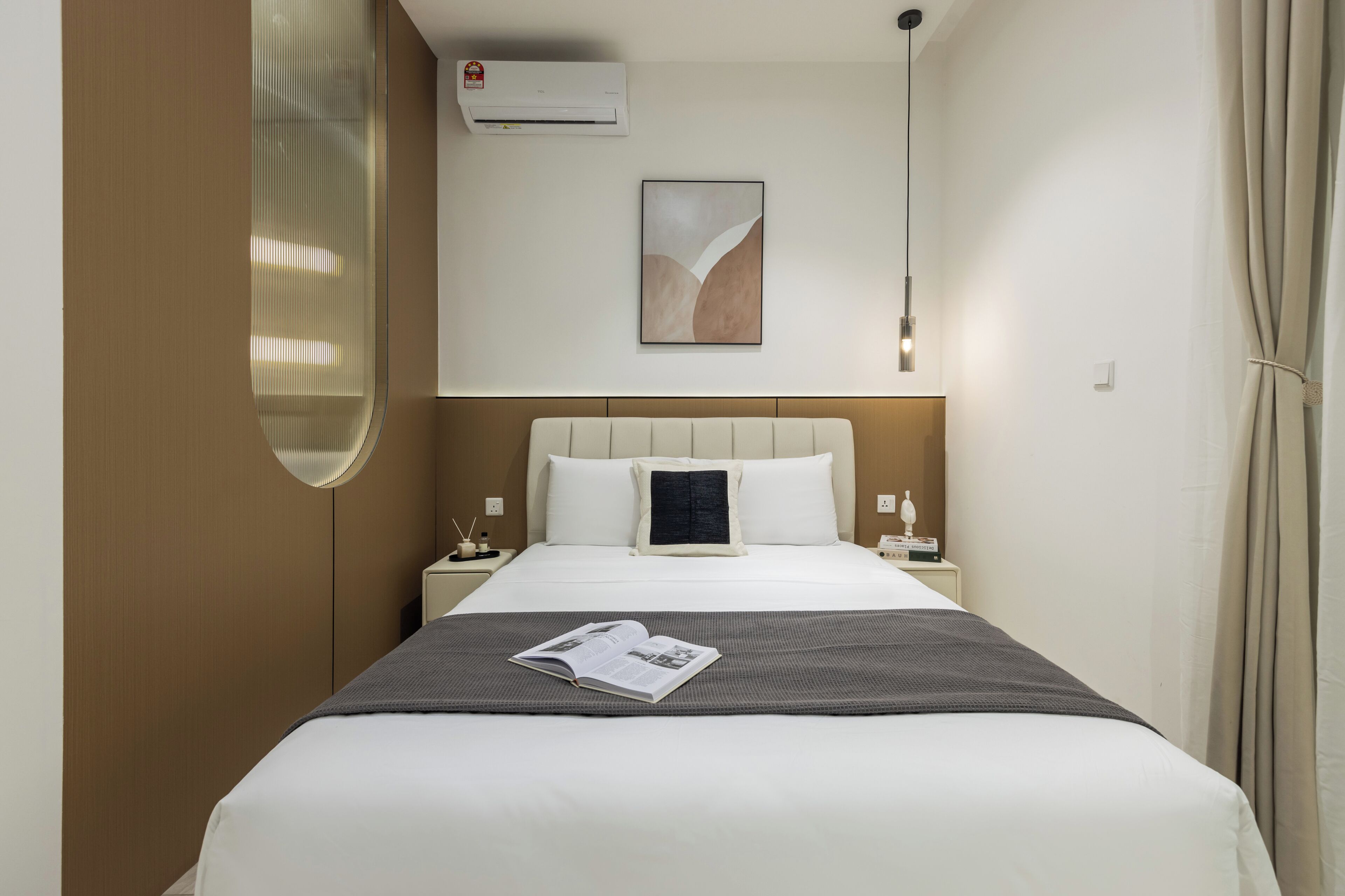 A hotel room with a bed, a bedside table, a nightstand, an air conditioning unit, a wall-mounted artwork, and a curtain.