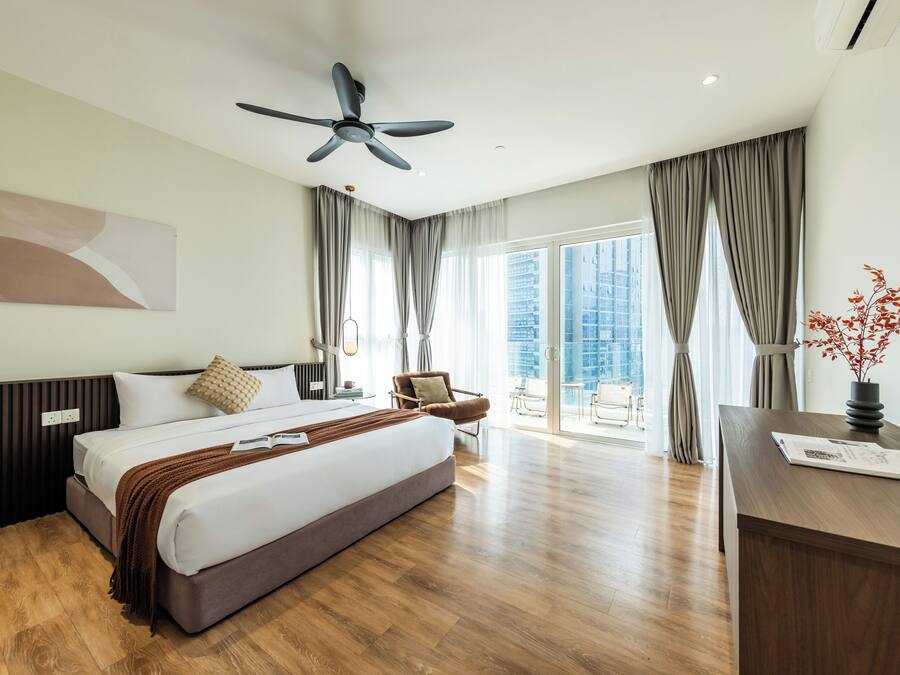 A modern hotel room with a large bed, a desk, a chair, a ceiling fan, and a view of a cityscape through the windows.