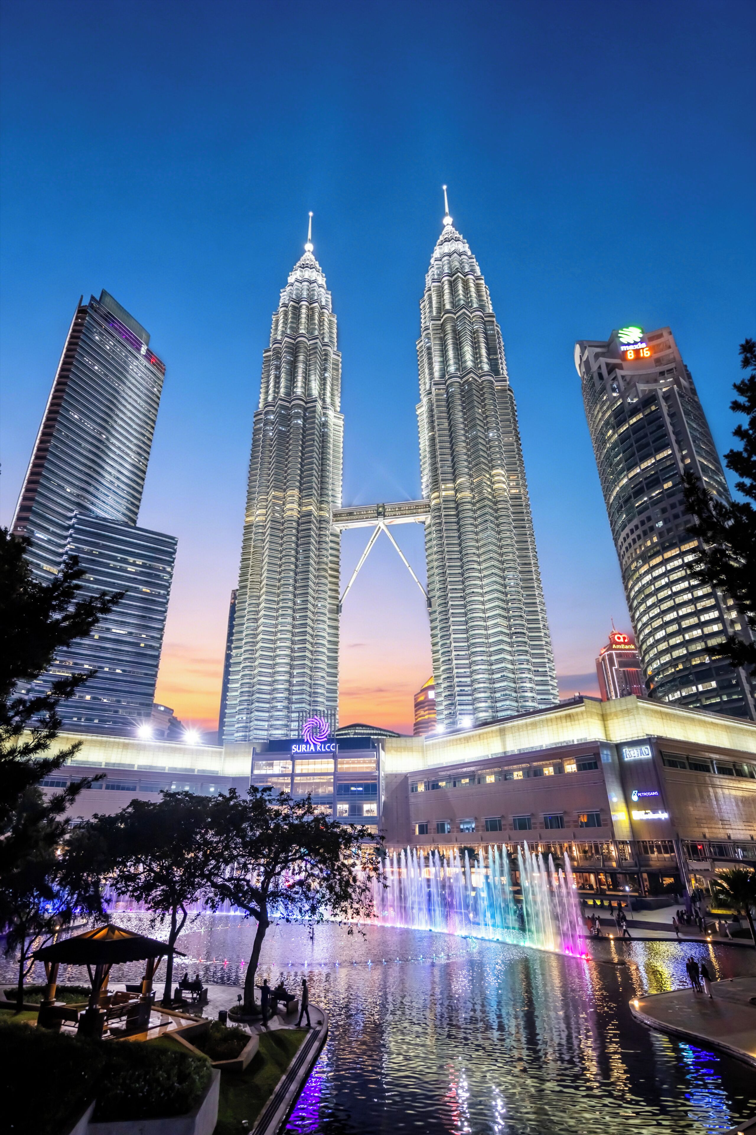 A cityscape with two prominent skyscrapers, a water feature with fountains, and surrounding modern buildings.