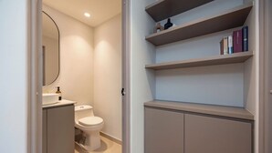 A modern bathroom with a toilet, a sink, and a mirror. There are shelves above the toilet with books and decorative items. - Live in Provenza Modern 1 BR   security (Medellín)