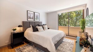 A bedroom with a large bed, a nightstand, a window with a view of trees, and framed pictures on the wall. - Live in Provenza Modern 1 BR   security (Medellín)