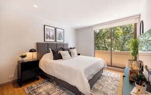 A bedroom with a large bed, a nightstand, a window with a view of trees, and framed pictures on the wall. - Live in Provenza Modern 1 BR   security (Medellín)