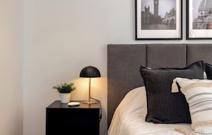 A neatly made bed with a grey headboard, a bedside table with a lamp, and two framed pictures on the wall. - Live in Provenza Modern 1 BR   security (Medellín)