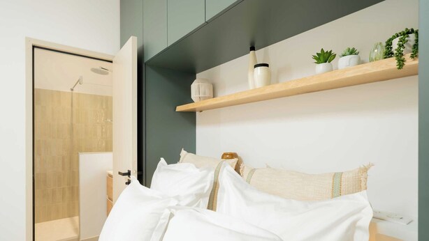A modern bedroom with a bed, a wooden door, a shelf with plants, and a wall-mounted light fixture.