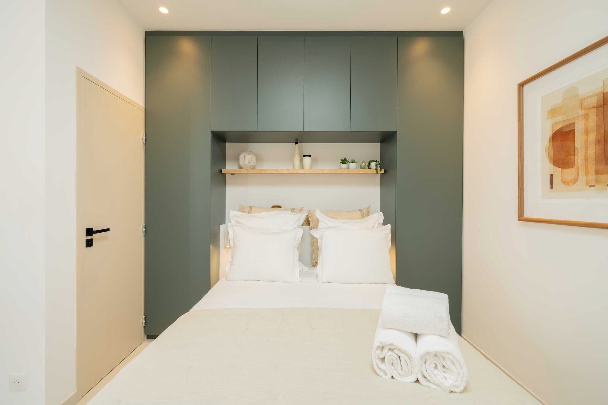 A hotel room with a bed, a wardrobe, a shelf with toiletries, and a framed picture on the wall.
