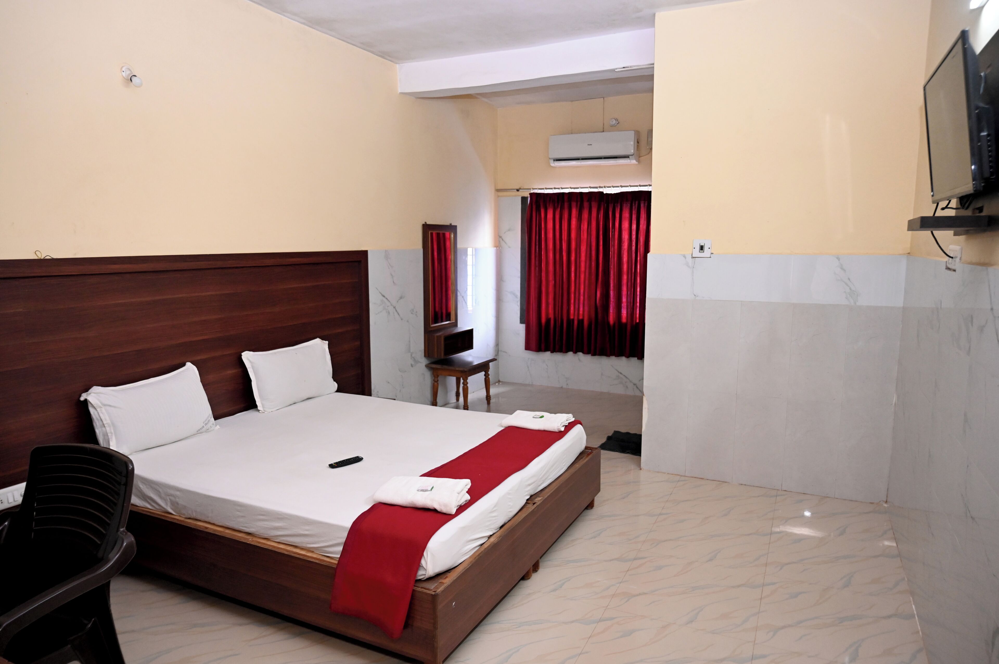 Une chambre d’hôtel avec un lit en bois, du linge de lit blanc, un plaid rouge, un téléviseur fixé au mur et un rideau rouge.