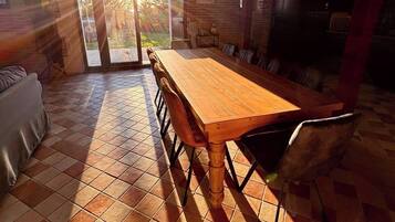 Zona pranzo con un grande tavolo di legno e sedie, pavimento piastrellato e vista attraverso una porta di vetro.