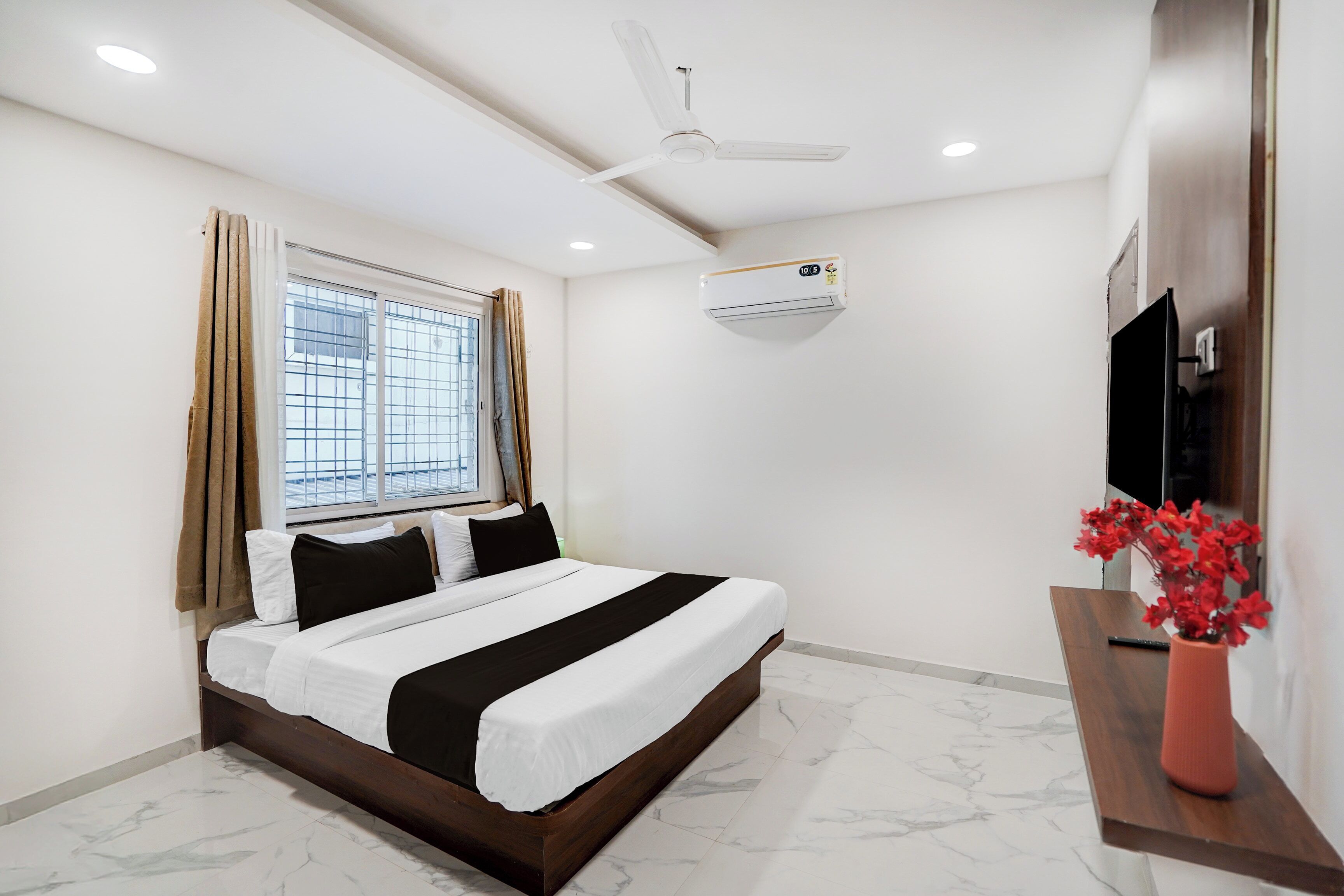 Habitación de hotel con cama, ventana con cortinas, ventilador de techo, unidad de aire acondicionado, televisor y un florero con flores.