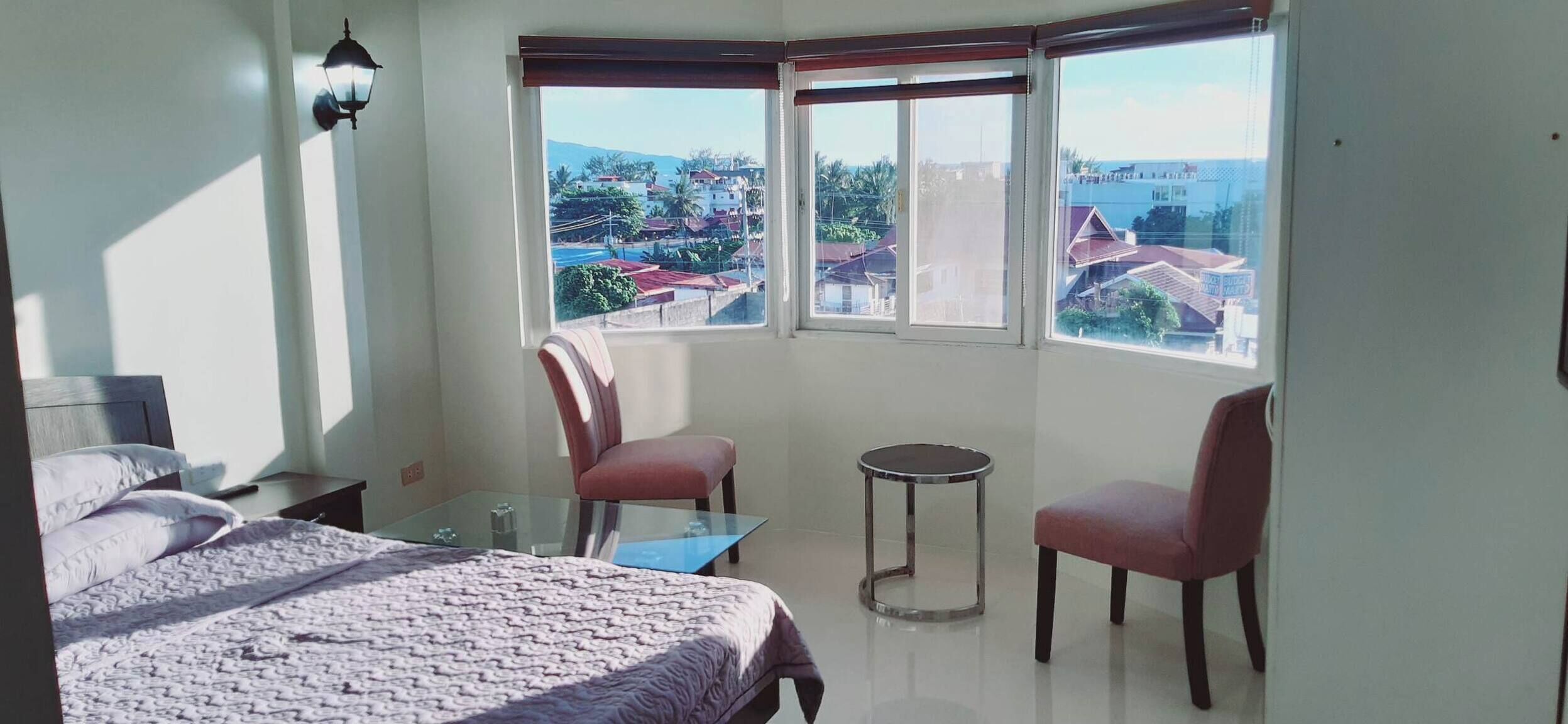 Un dormitorio con cama, dos sillas, una mesita y vistas a una zona residencial a través de amplias ventanas.