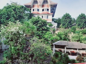 Ein mehrstöckiges Gebäude mit markanter Architektur, umgeben von üppiger Vegetation und einem Gebäude mit einem Strohdach.