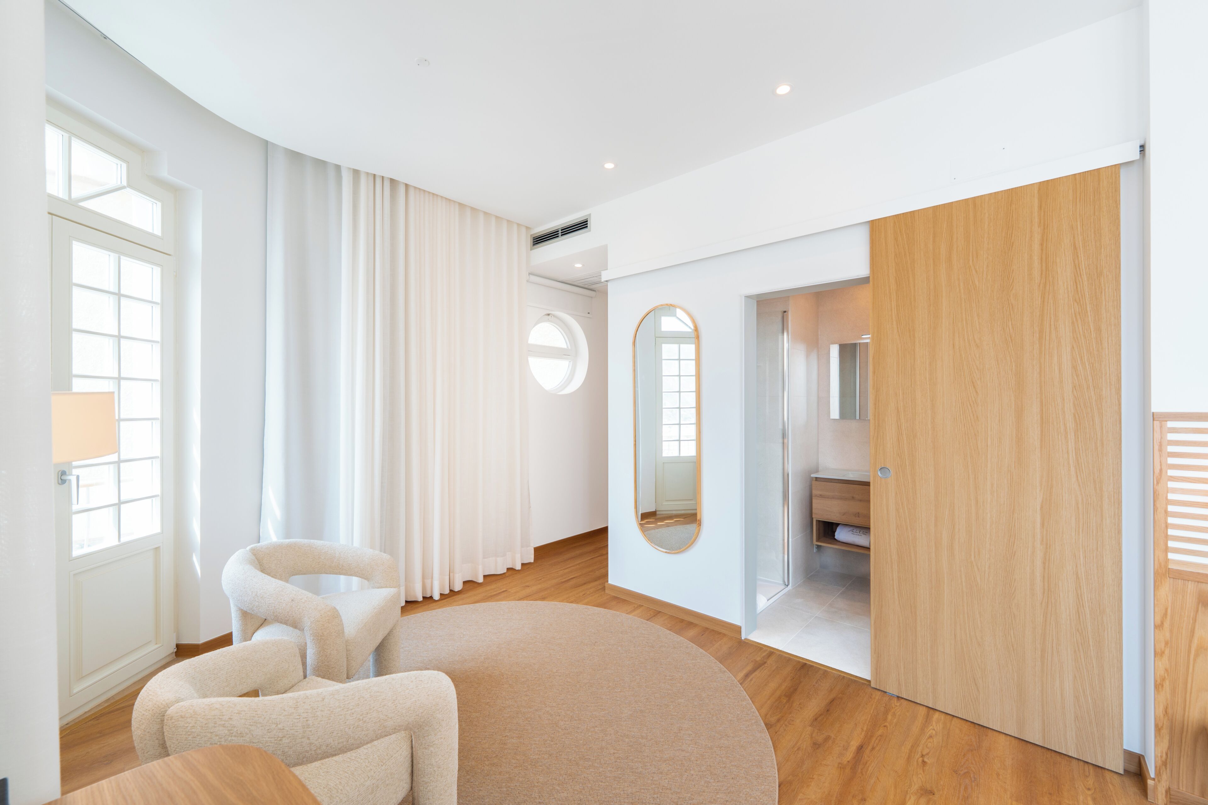 A modern hotel room with a large window, a round mirror, a wooden door, and a seating area with a round rug.