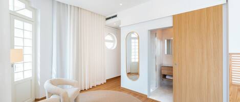A modern hotel room with a large window, a round mirror, a wooden door, and a seating area with a round rug.