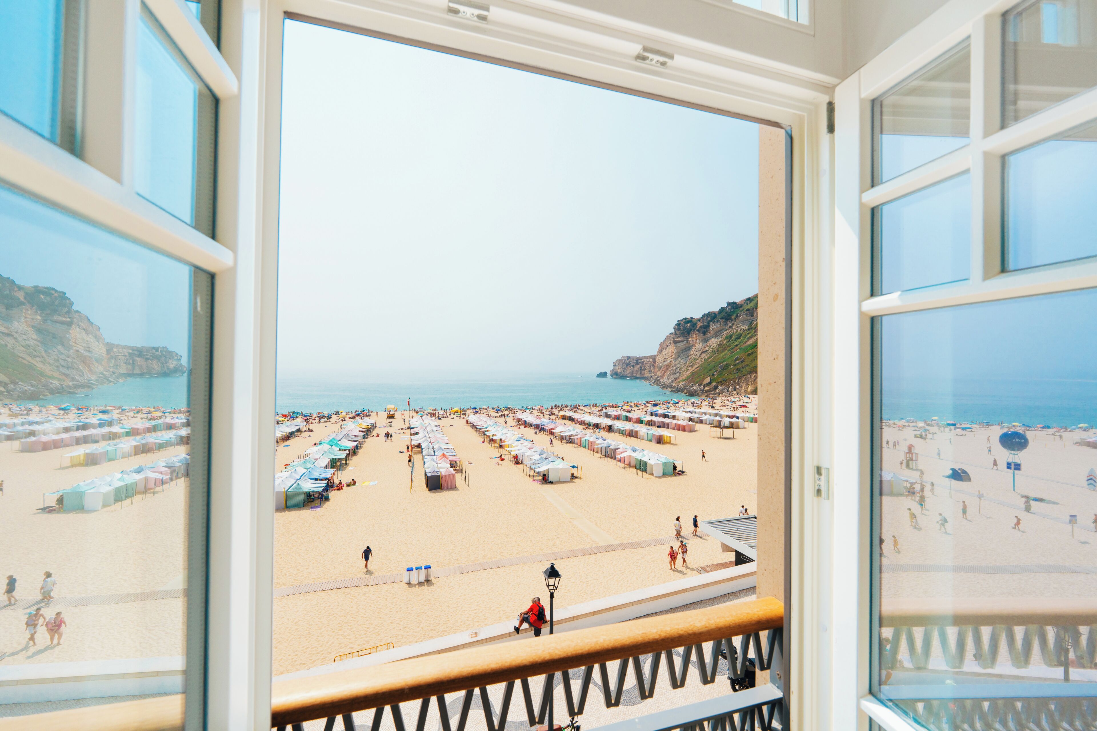 A beach view from a window with a railing, featuring a sandy beach with numerous tents and people.