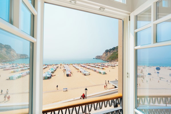 A beach view from a window with a railing, featuring a sandy beach with numerous tents and people.