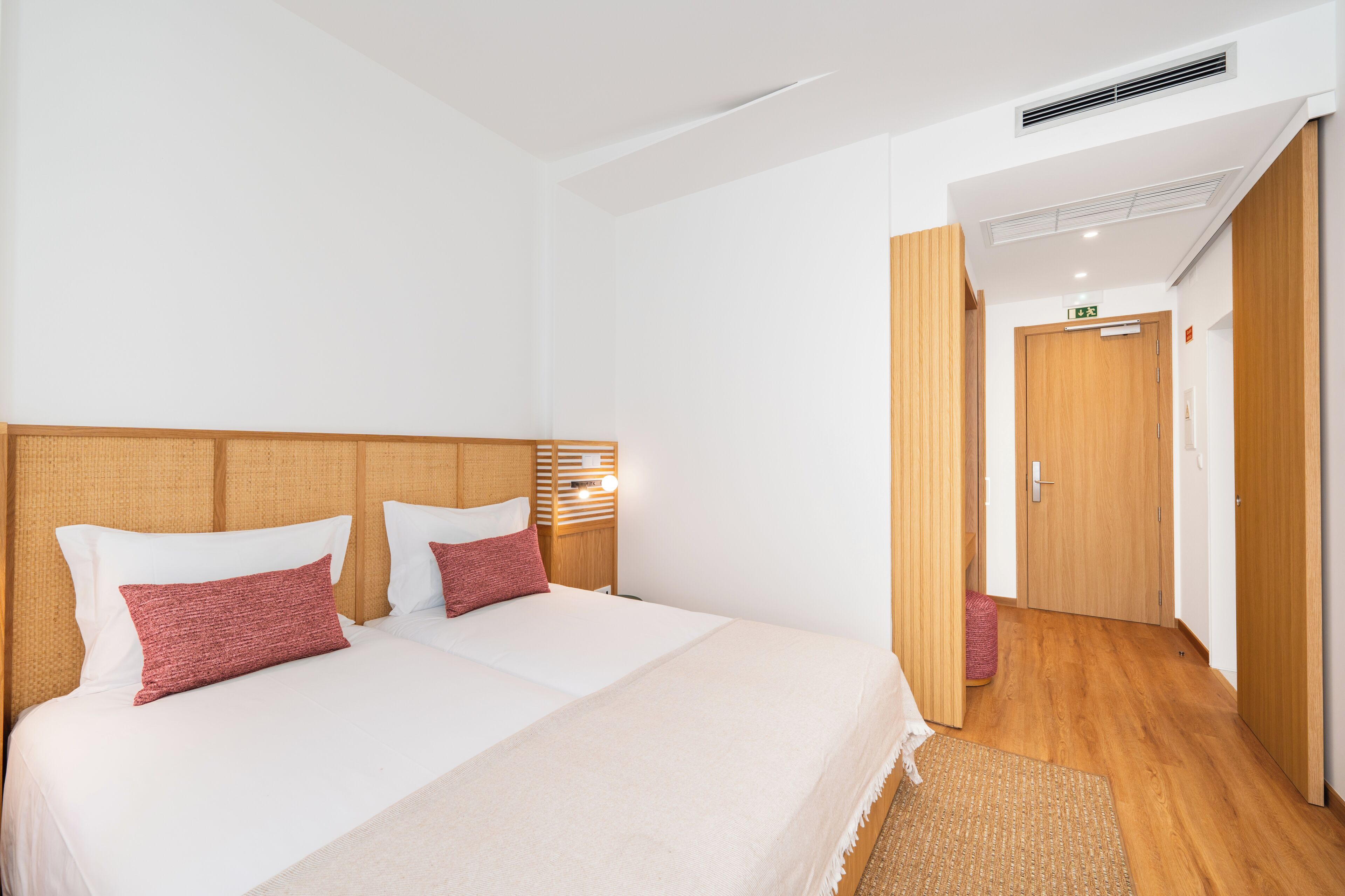 Habitación de hotel con una cama grande, cabecero, mesita de noche y lámpara de pared, además de una puerta que da a otra habitación.